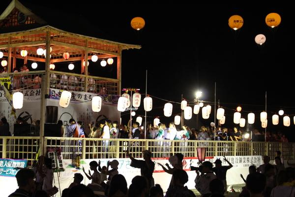 ライトアップされたやぐらの周りを踊る、沢山の人々の写真