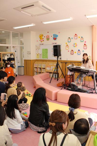 西田 夫佐さんがキーボートを弾きながらマイクで歌を歌いそれを観覧している親子たちの写真