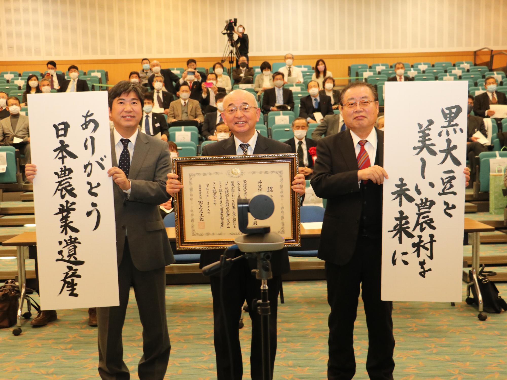 市長が認定証を持ち、両隣の方が「ありがとう日本農業遺産」「黒大豆を美しい農村を未来に」と書かれた書を持っている。