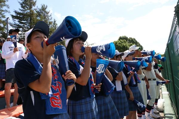 青いプラスチックのメガホンを口元に応援をしている女子生徒と、ユニフォームを着て応援している男子生徒の写真