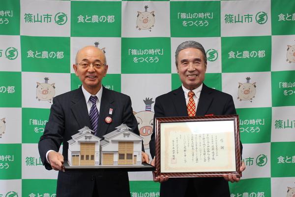 市長が山倉2棟の模型をもち、スーツ姿の男性が額に入った賞状を持っている写真