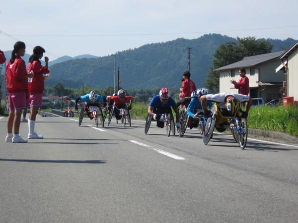 6人のランナー達がマラソンコースで競り合いながら車いすをこいでいる写真