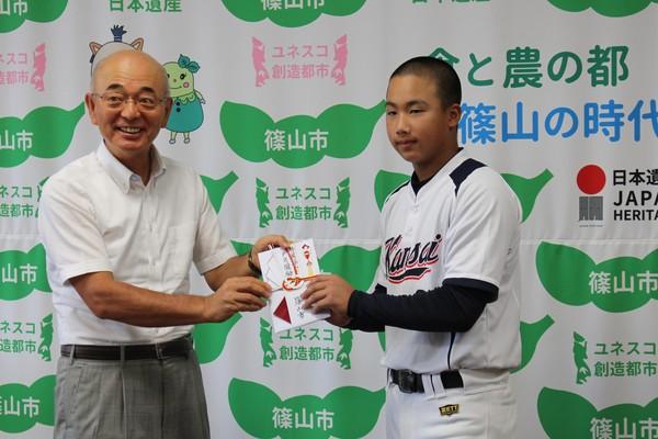 野球のユニホームを着た少年野球関西選抜チームの代表として全国大会に出場する萱野くんに市長がのし袋を手渡している写真