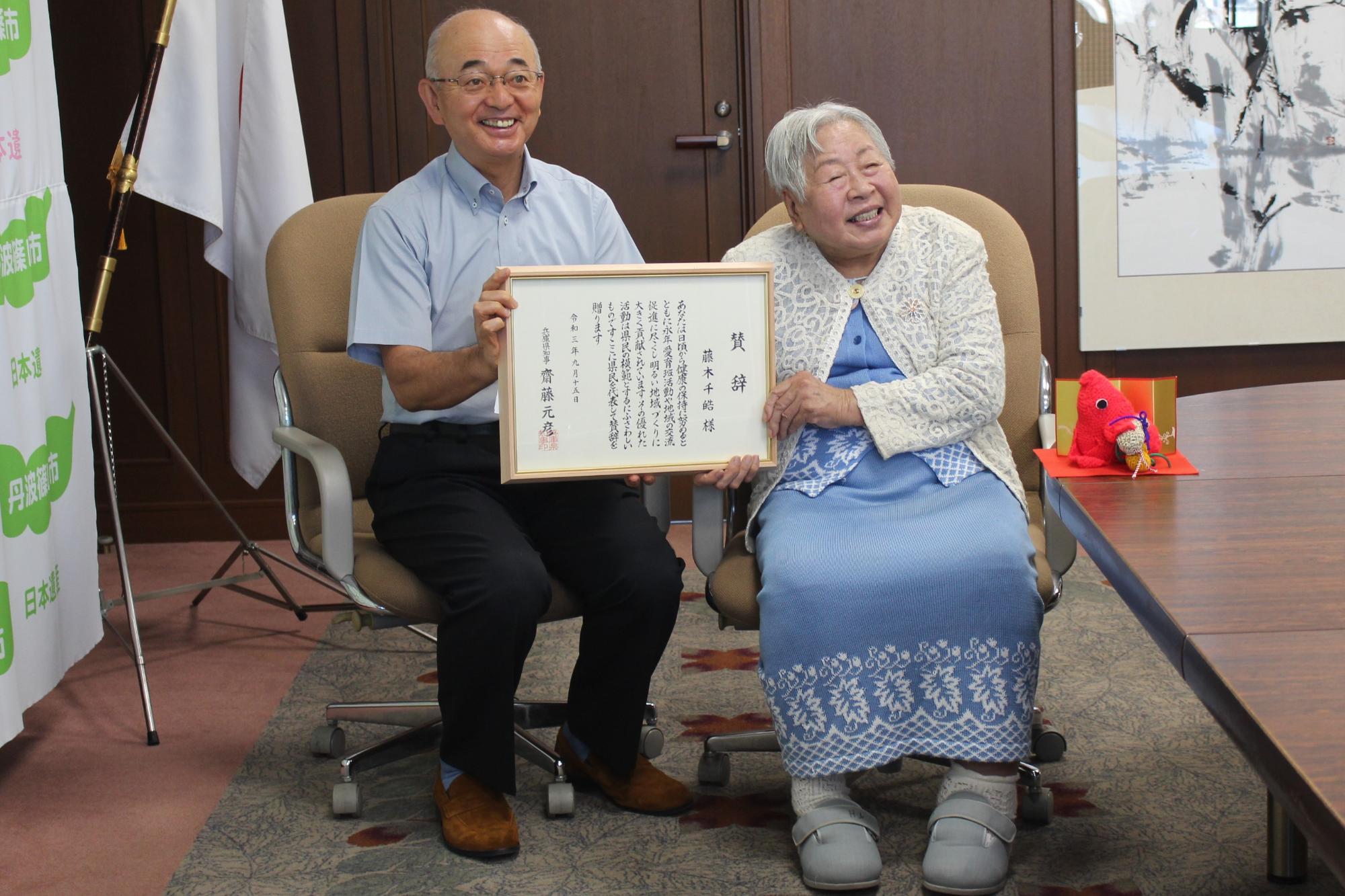 椅子に座って市長と女性が一緒に表彰状を持っている。