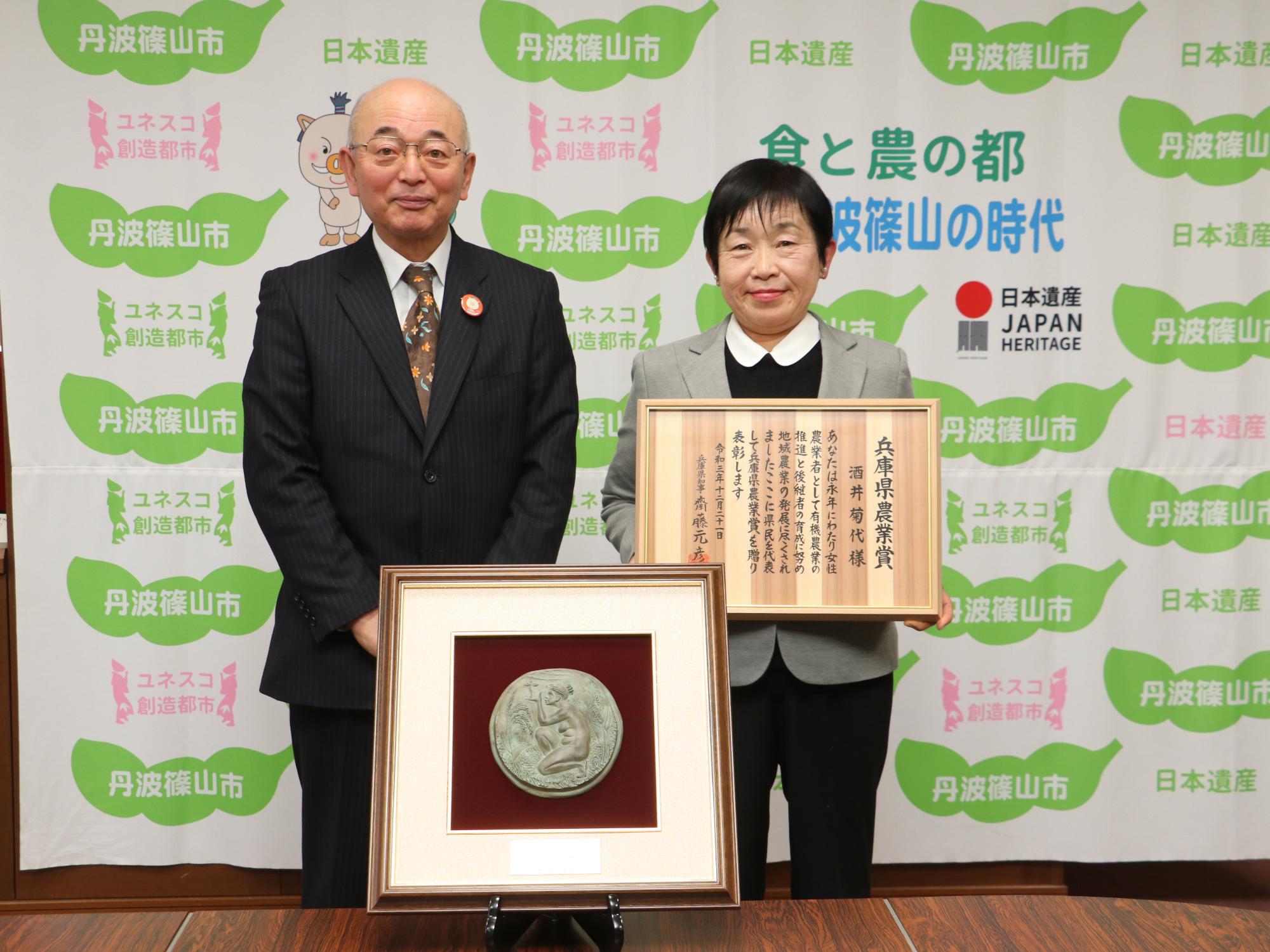 机に焼き物の盾があり、その後ろに表彰状を持った女性と市長が立っている