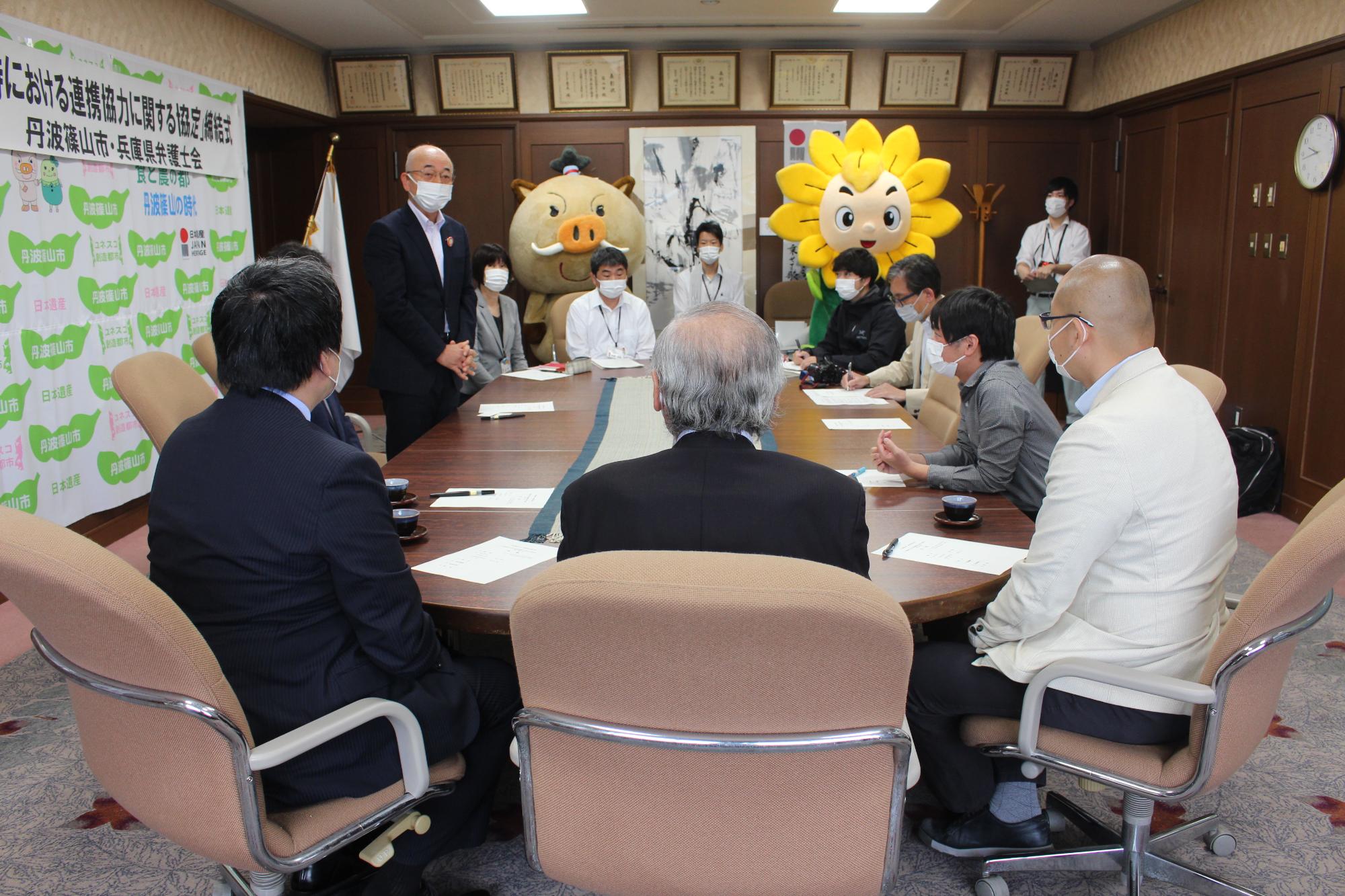 応接室で市長が立って説明をされている。奥にはマスコットキャラクターが並んで立っている。