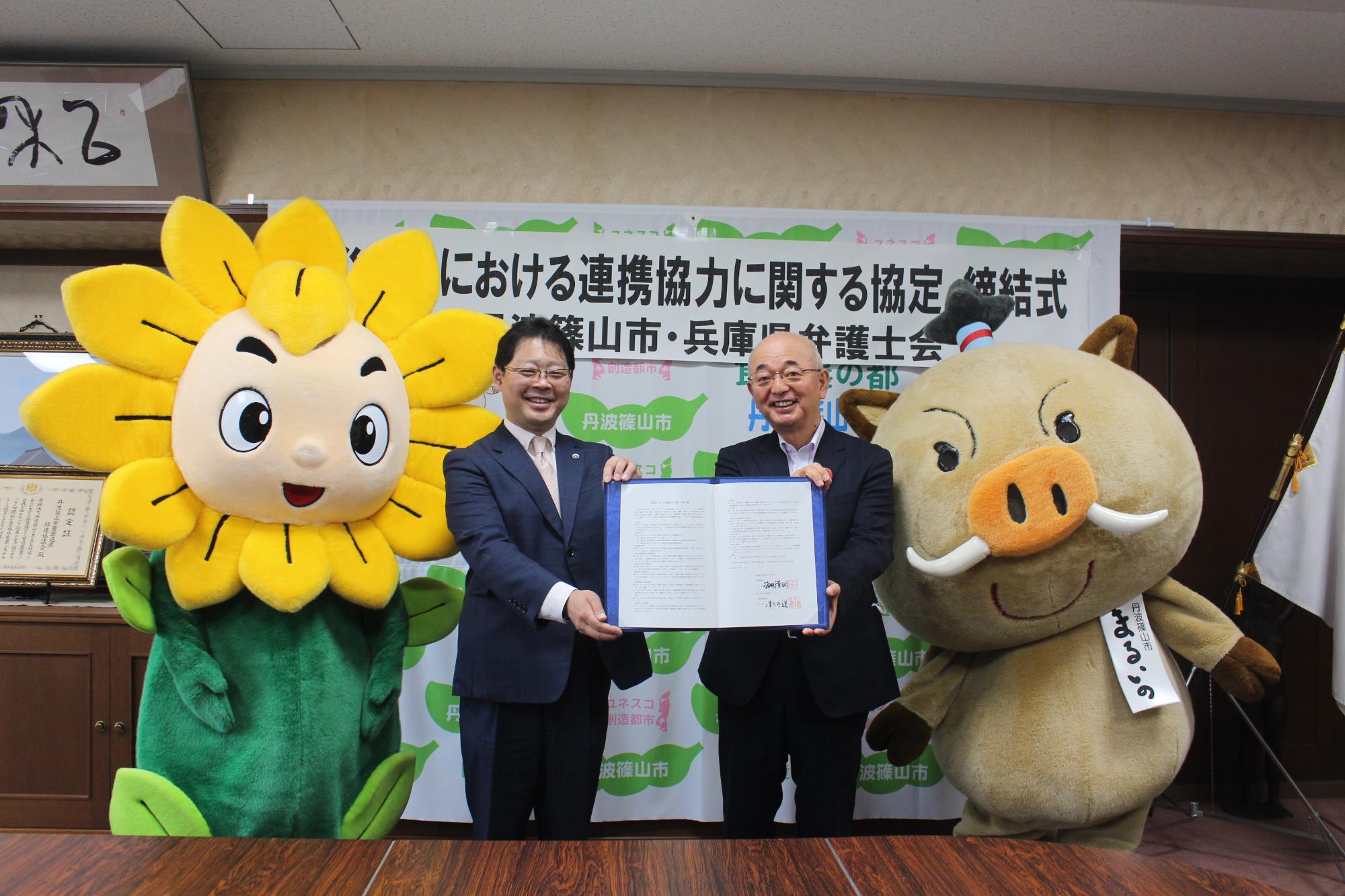 市長と兵庫県弁護士会会長が、協定書を一緒に持っている。隣にはマスコットキャラクターのまるいのとヒマリオンが並んでいる。