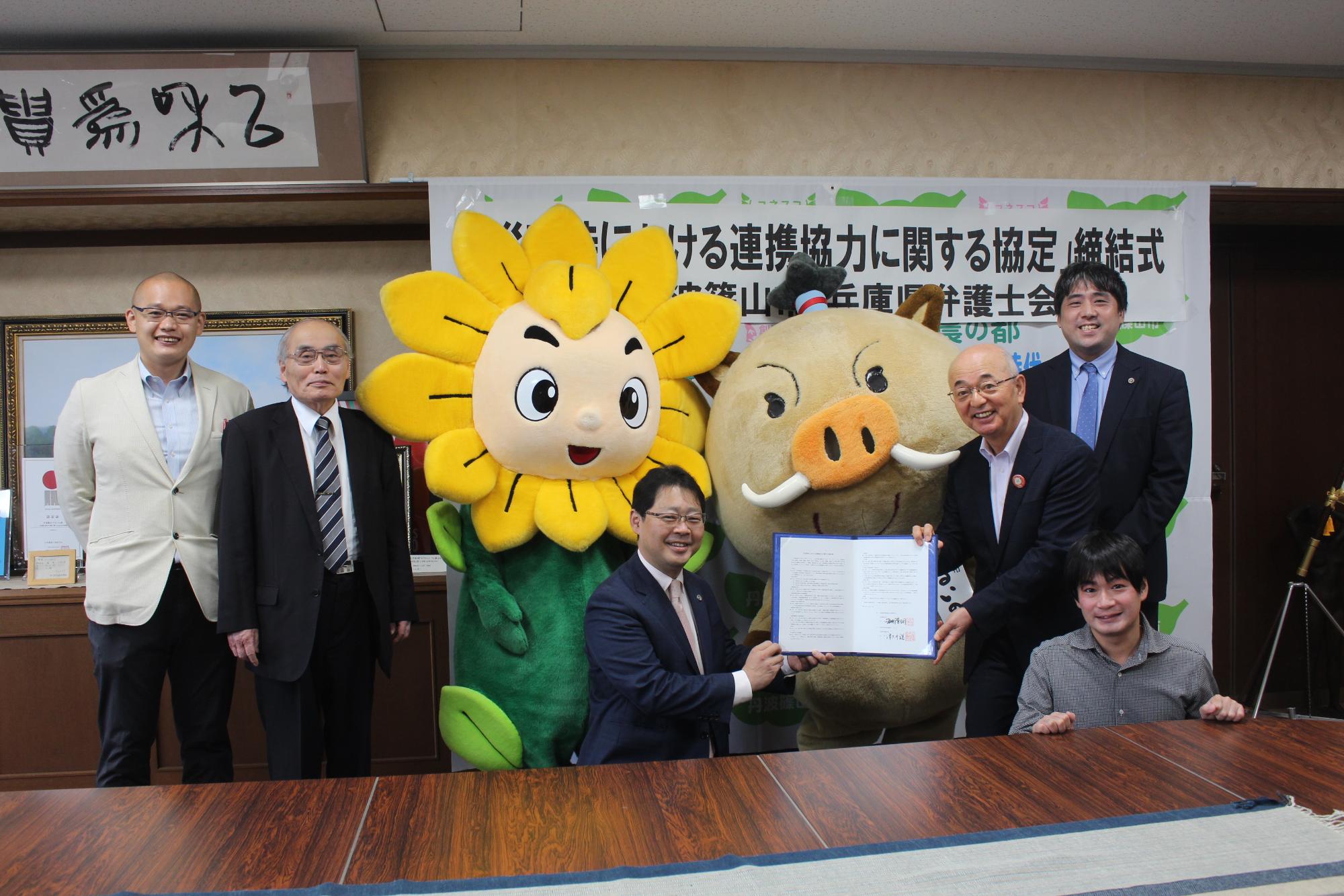 市長と兵庫県弁護士会の会長が一緒に協定書を持っていて、まわりには弁護士協会の方や関係者、マスコットキャラクターのまるいのとヒマリオン。