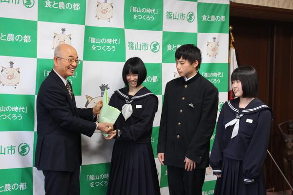 生徒会役員3名のうち、女子生徒が集まった支援金を市長に手渡ししており、他の2名の生徒が渡す様子を見ている様子の写真
