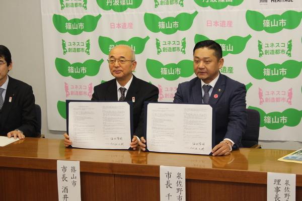 泉佐野市の千代松市長と丹波篠山市の市長が、協定書にサインした証書を机の上に広げて開示している写真