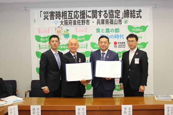 左端が、篠山青年会議所 青木理事長、丹波篠山市の市長、泉佐野市の千代松市長、右端が泉佐野青年会議所 宮内理事長が写っており、市長がそれぞれ協定書を手に持って開示している写真