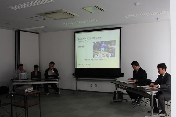 職員が席に座ってスクリーンを使いながら中間報告をしている写真