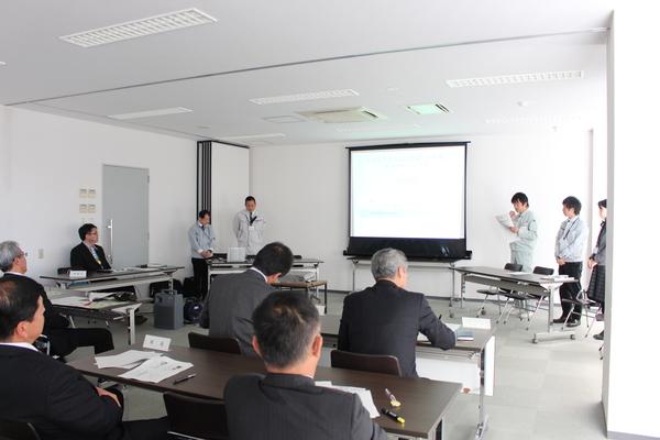 研究職員がみんなで立ち代表者の男性がスクリーンを使って発表している写真