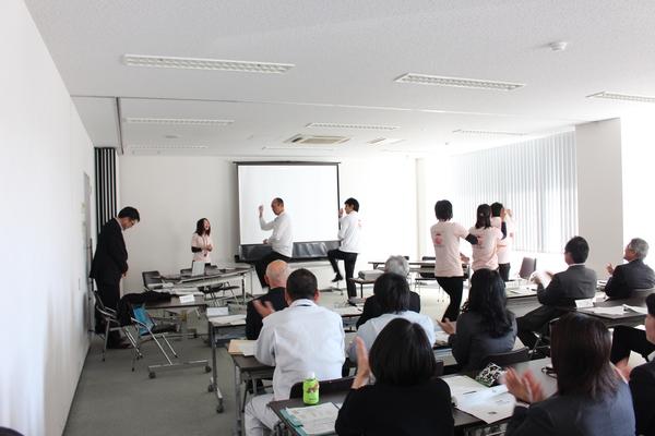 職員たちがスクリーンの前でデカンショ踊りを披露している写真