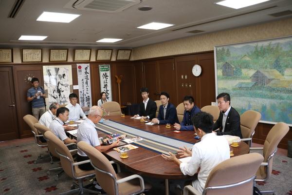 楕円形のテーブルに10名ほどが座り中心の男性の話を聞いている写真