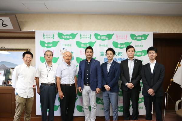 篠山市の文字が入った壁の前で7名の男性が並んで記念撮影をしている写真