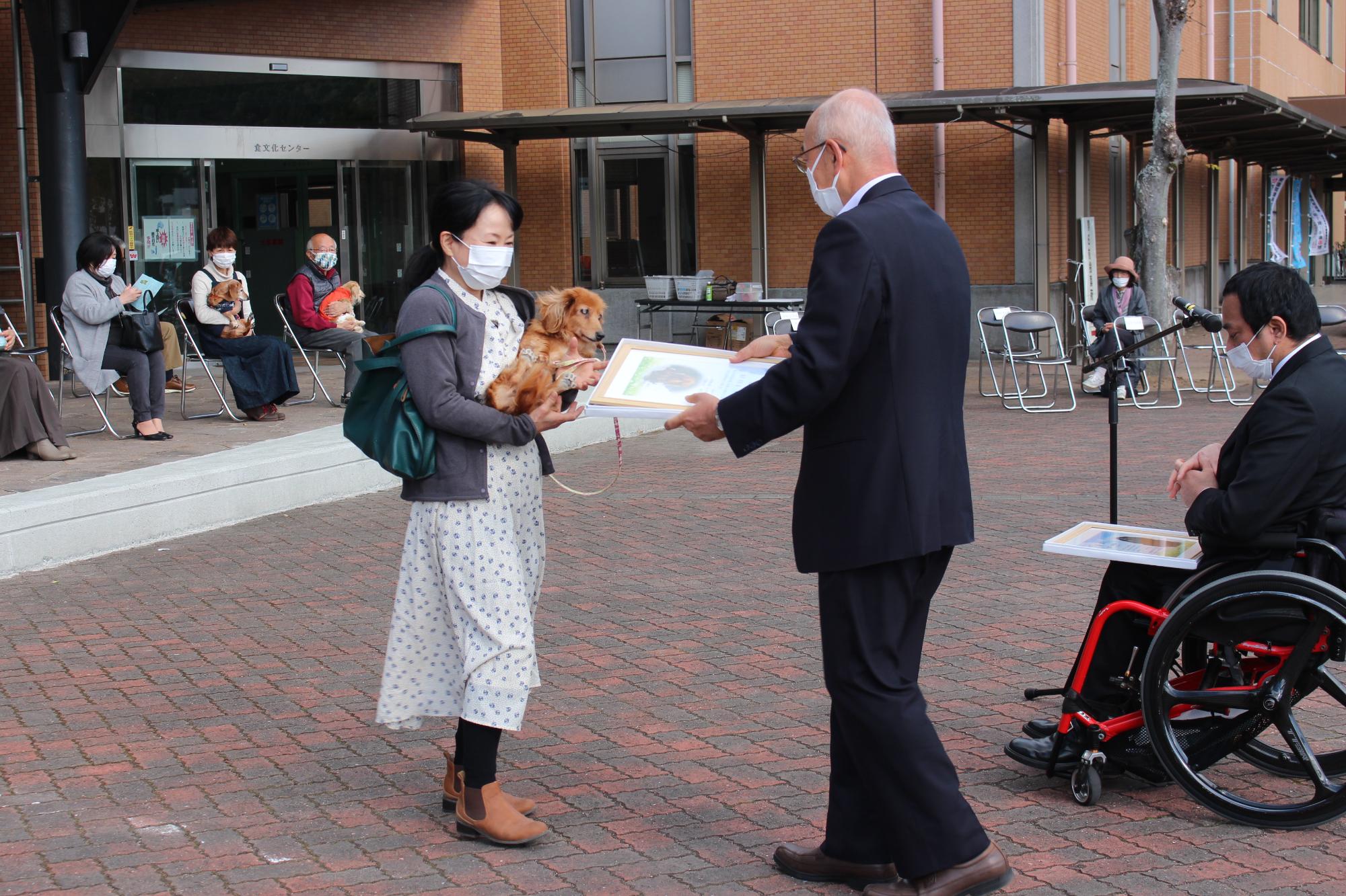 長寿犬を抱いた女性が、市長から表彰状を受け取ろうとしている。