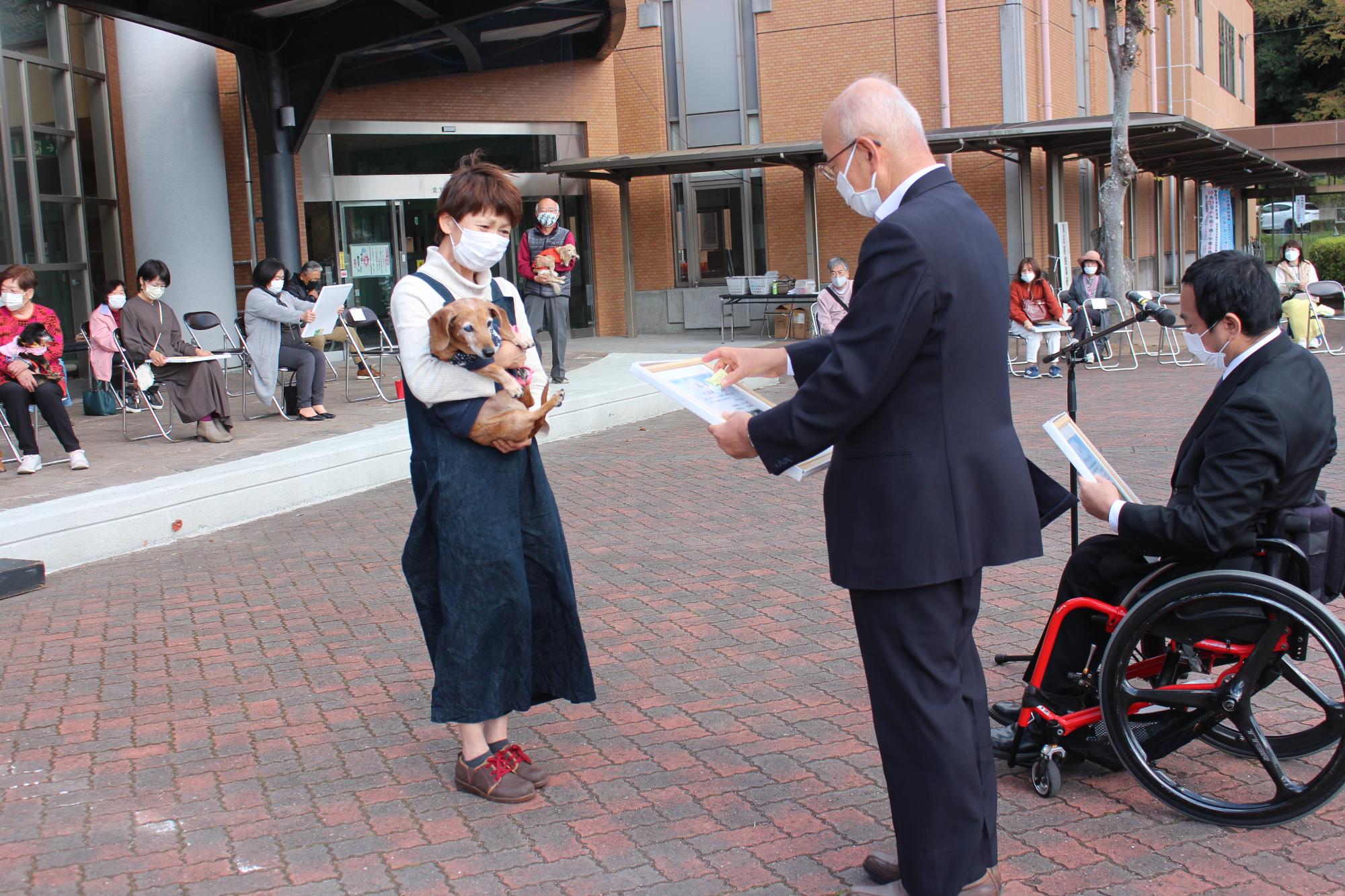 長寿犬を抱いた女性が、市長から表彰状を受け取ろうとしている。