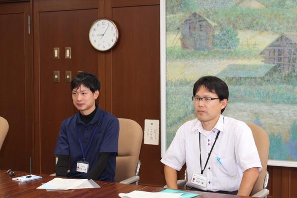 机の左側にショートカットの安井主事、隣に眼鏡をかけた田中係長が座っている写真