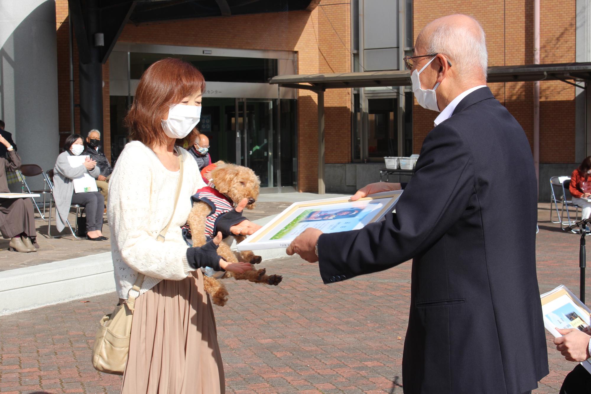 長寿犬を抱いた女性が、市長から表彰状を受け取ろうとしている。