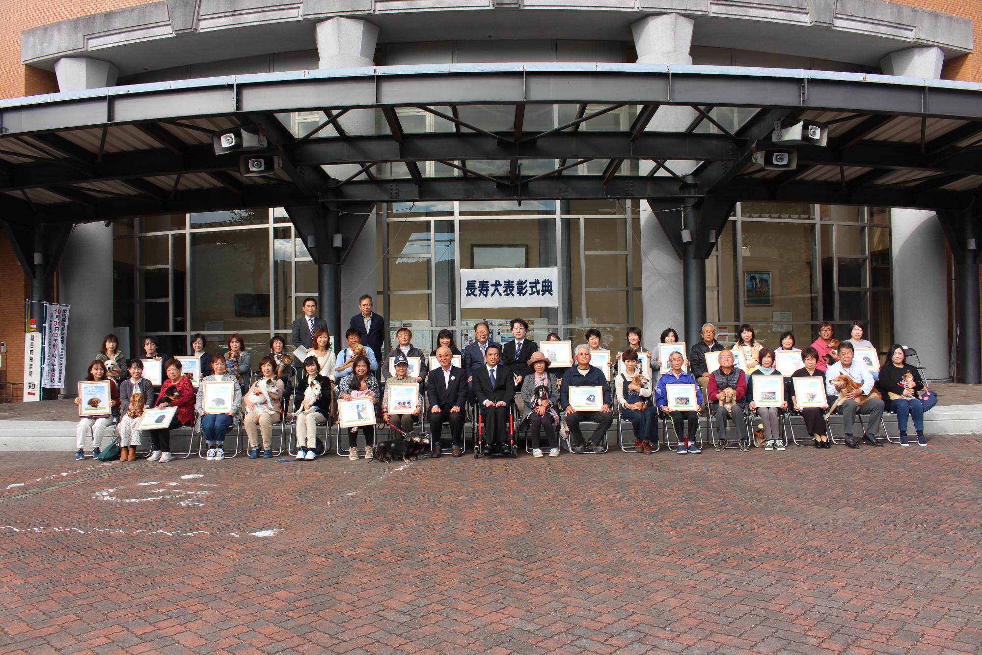 市長と獣医師会の皆さん、長寿犬と飼主の皆さんがパイプ椅子に座って記念撮影。