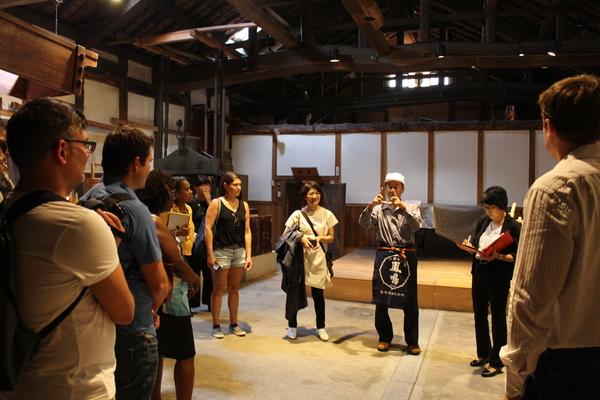 世界各国から研修に来た外国人の男女が、腰にエプロンをつけて話す男性に周りに集まっている写真