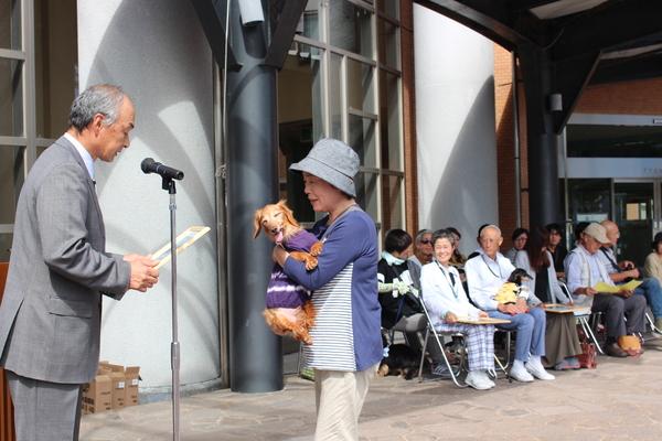 洋服を着た長寿犬が帽子を被っている飼い主の方に抱っこされ表彰されている写真
