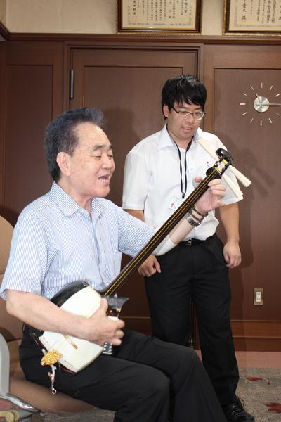 山口先生が歌いながら三味線を弾いていて、その後ろでメガネをかけた男性職員が歌っている写真