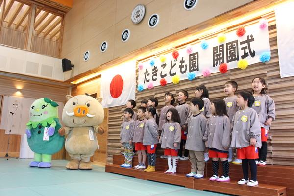 園児が開園式でマスコット人形と一緒に歌を歌っている写真
