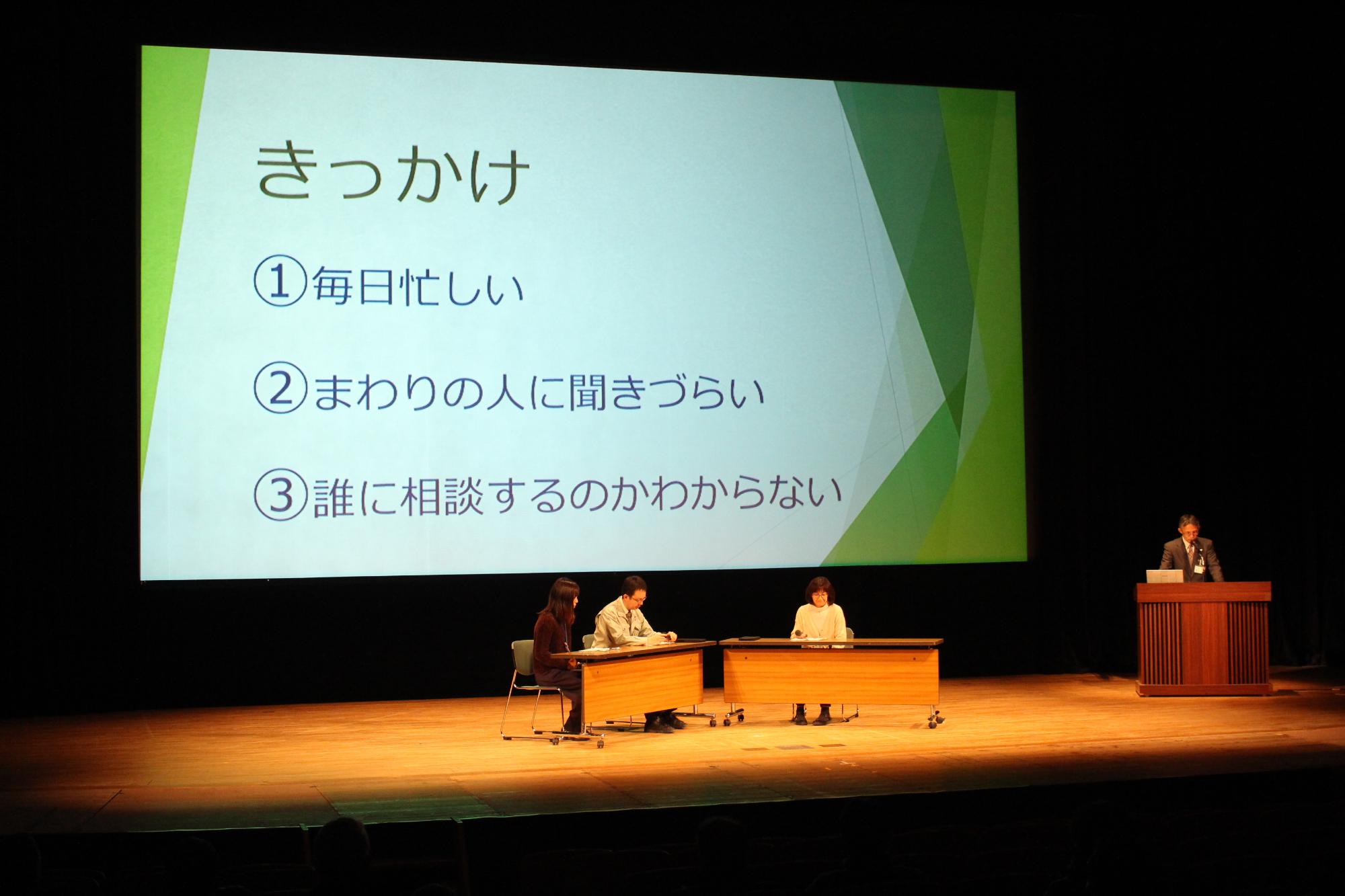 毎日忙しい、誰に相談すればよいかわからないなどお助け掲示板を作成するきっかけをスライドで写し、イスにすわった3名と、立って説明をする総務課の職員