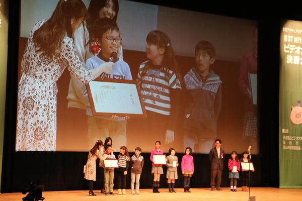 ペイズリー柄のワンピースを着た女性が、受賞を受けた小学生チームの男の子にマイクを向けており、その映像が後ろの大きいスクリーンに映し出されている様子の写真