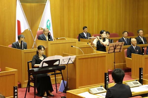 議場にてフルート奏者の松村 京子さんと電子ピアノ奏者の井本 裕美さんが演奏しており、議員の方々が、議員席に座って演奏に聞き入っている様子の写真