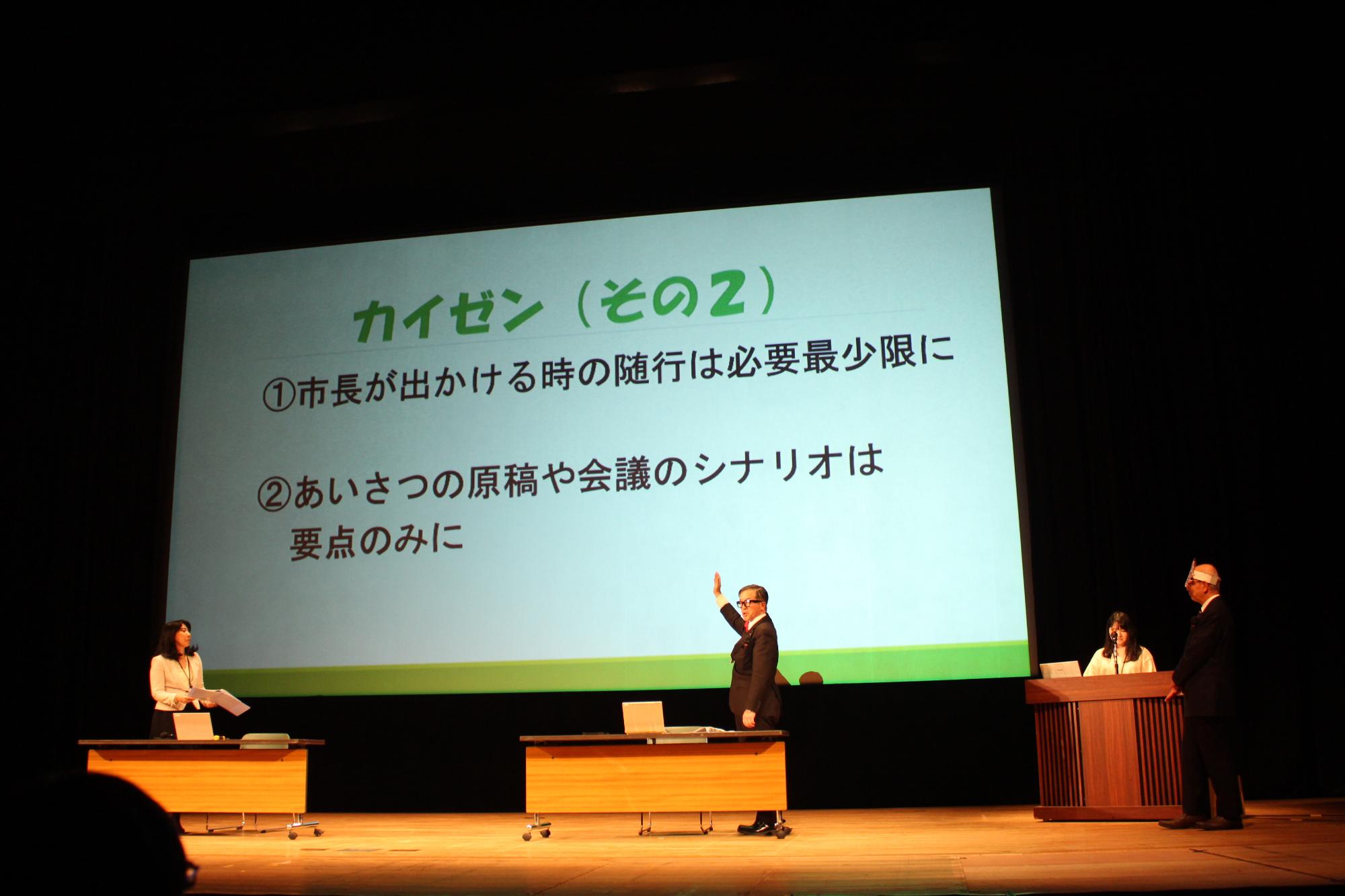「カイゼン（その2）1.市長が出かける時の随行は必要最小限に 2.あいさつの原稿や会議のシナリオは要点のみに」と書かれたスライドを指し、説明する秘書課長