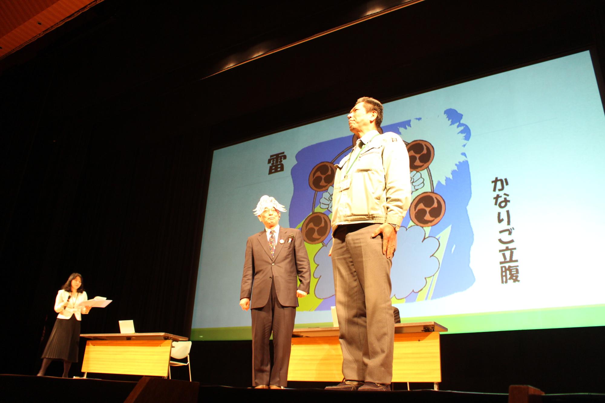 市長のお天気予報で、前のスクリーンに雷さんの市長が映し出され、説明をする秘書と、助っ人の怒られ役の職員とその横に立つ市長の写真