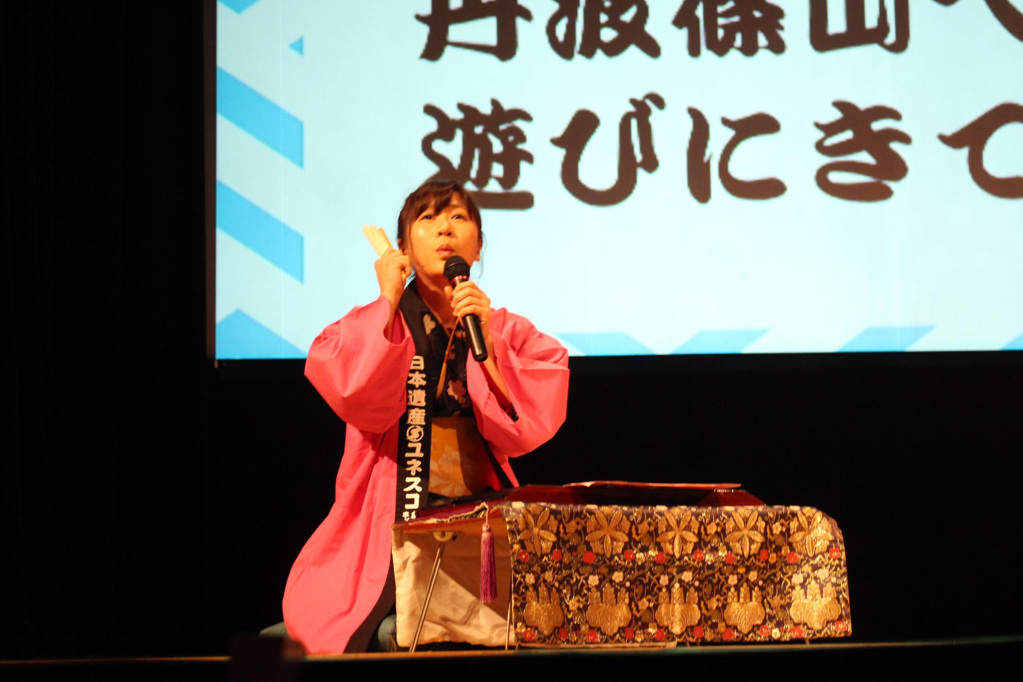 ピンクのハッピを着た女性職員が閉じた扇子とマイクを持ち、正座をして話している写真