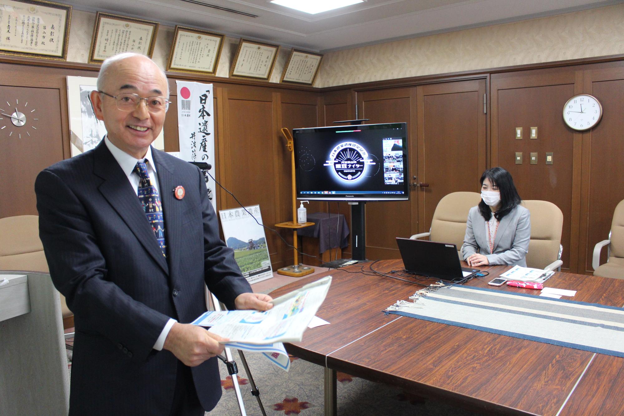 市長が資料を手に持ち、オンラインで中学生のこどもたちと交流している。奥に大きなモニターがあり、傍の机で女性職員がパソコン操作をしている