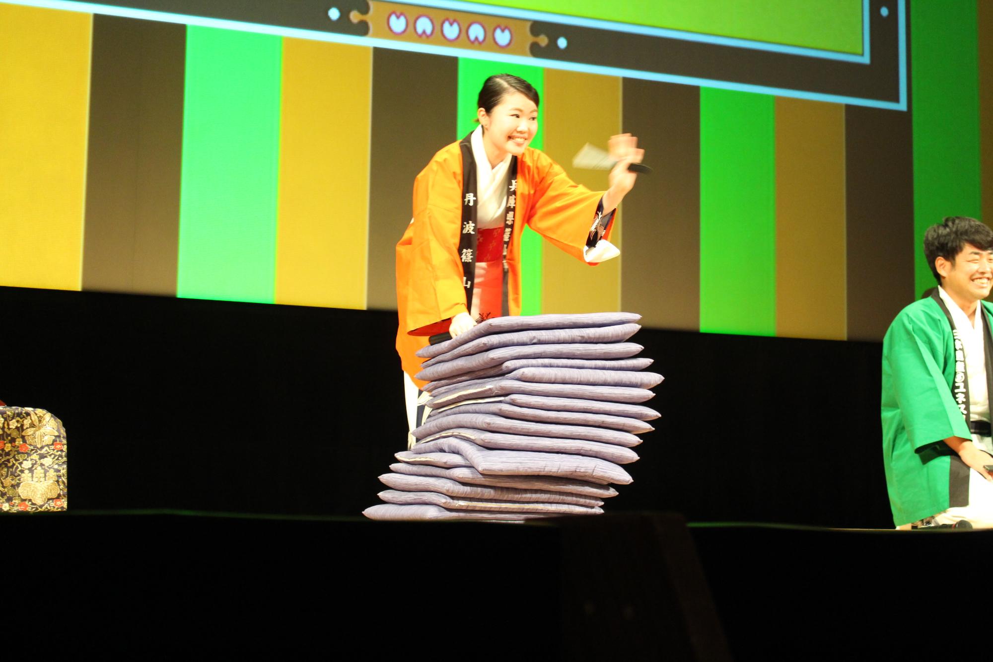 オレンジのハッピを着た女性職員がたくさん置かれた座布団を倒れないように右手で支え、閉じた扇子をもった左手を客席に向かってふっている写真