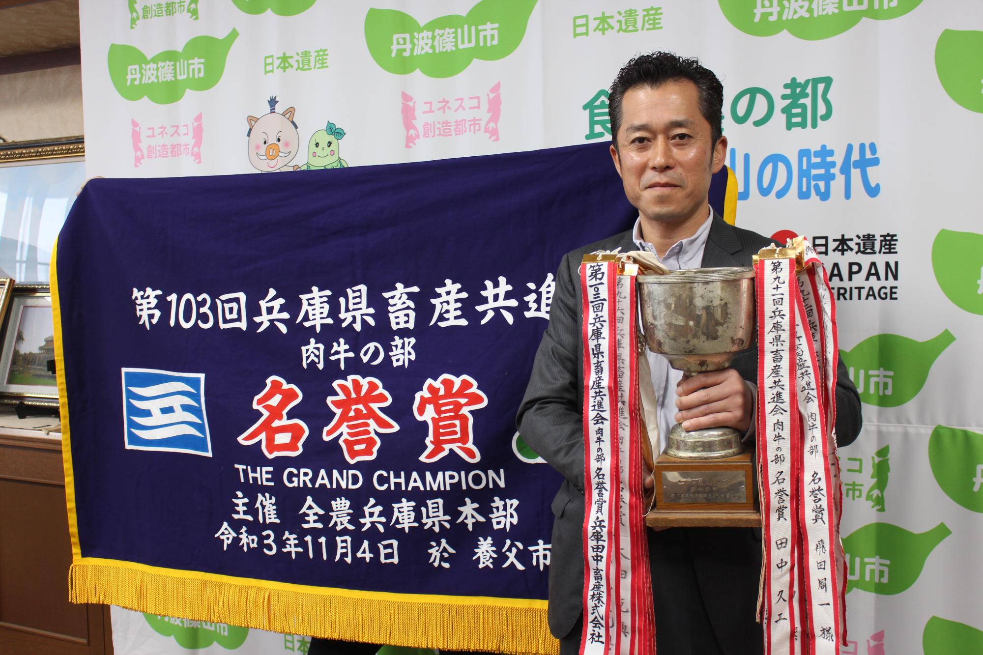 名誉賞と書かれた幕の前に、受賞された男性がカップを持って記念撮影