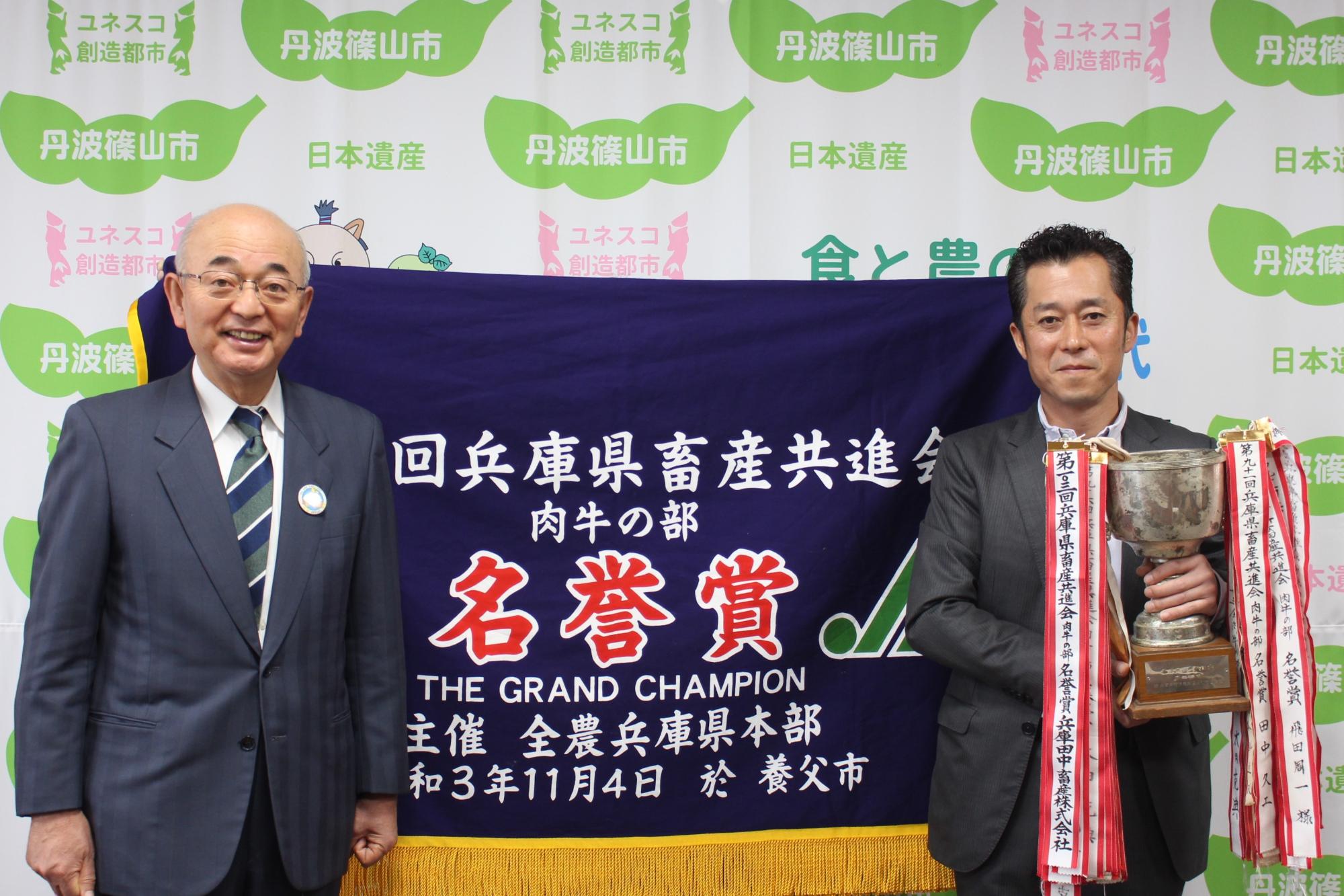 名誉賞と書かれた幕の前で、市長とカップを持った受賞者が記念撮影