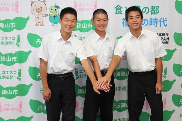 篠山市と書かれた壁の前で3人の中学生が中央で手を重ねている写真