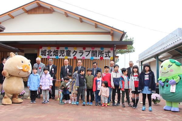 城北畑児童クラブ開所式と書かれた、建物の前に並ぶ子供たちと、ゆるキャラとスーツを着た関係者の写真