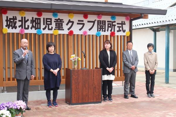 マイクを持って話をしている男性と、その横に立つ4名の男女の写真