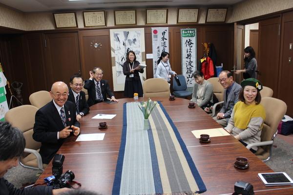 市長室にて、緑色に黄色と白の花が飾られているベレー帽をかぶった森田 まりこさんが市長の前に座っており、市長室にいる関係者が全員笑顔になっている様子の写真