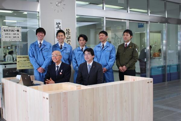 市長が設置の終わったカウンターに座り、その後ろに篠山産高土木科3年生が立って写っている写真
