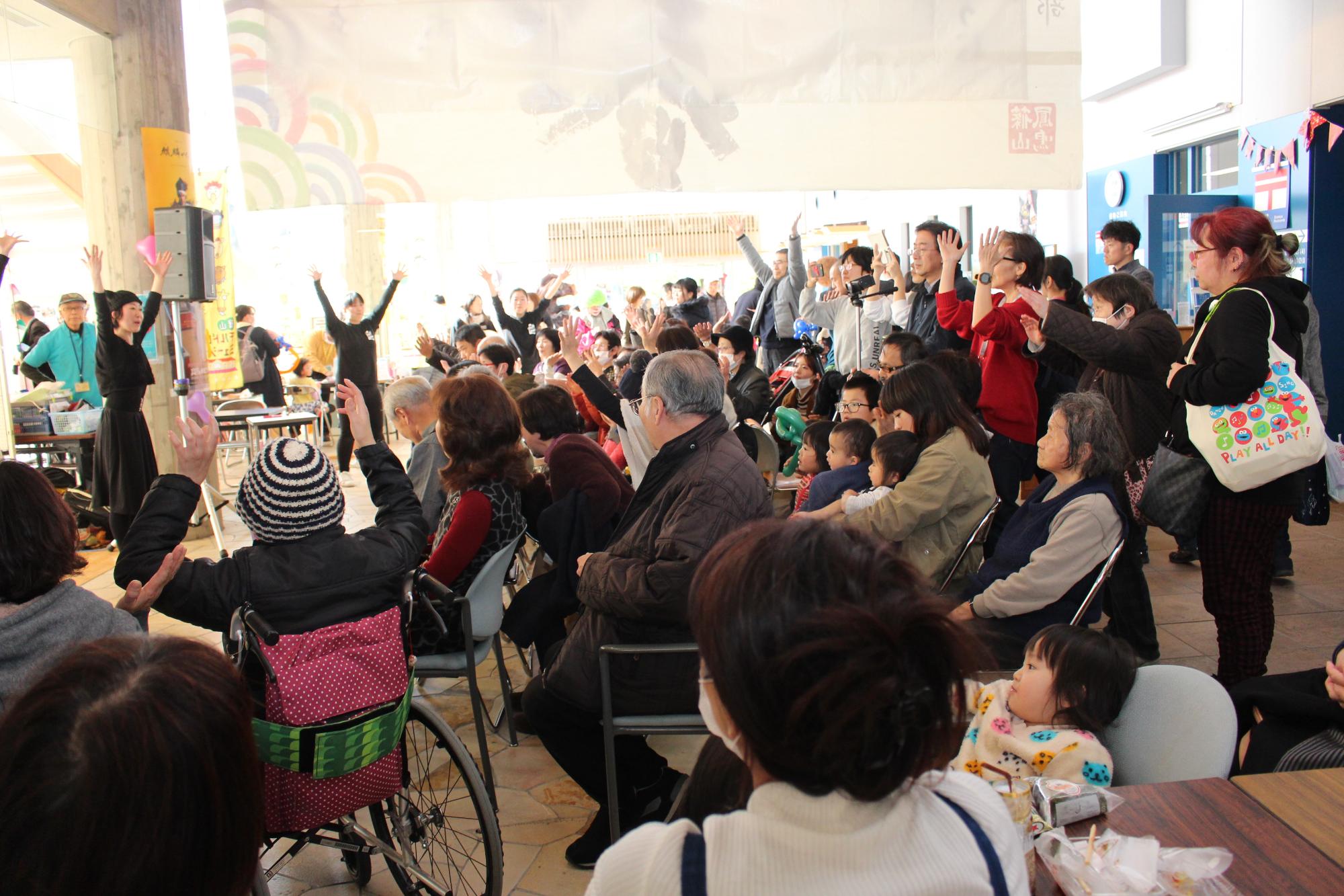 前で両手をあげて踊っている黒い服を着た女性たちと、そのまねをして両手をあげているたくさんの方たちの写真