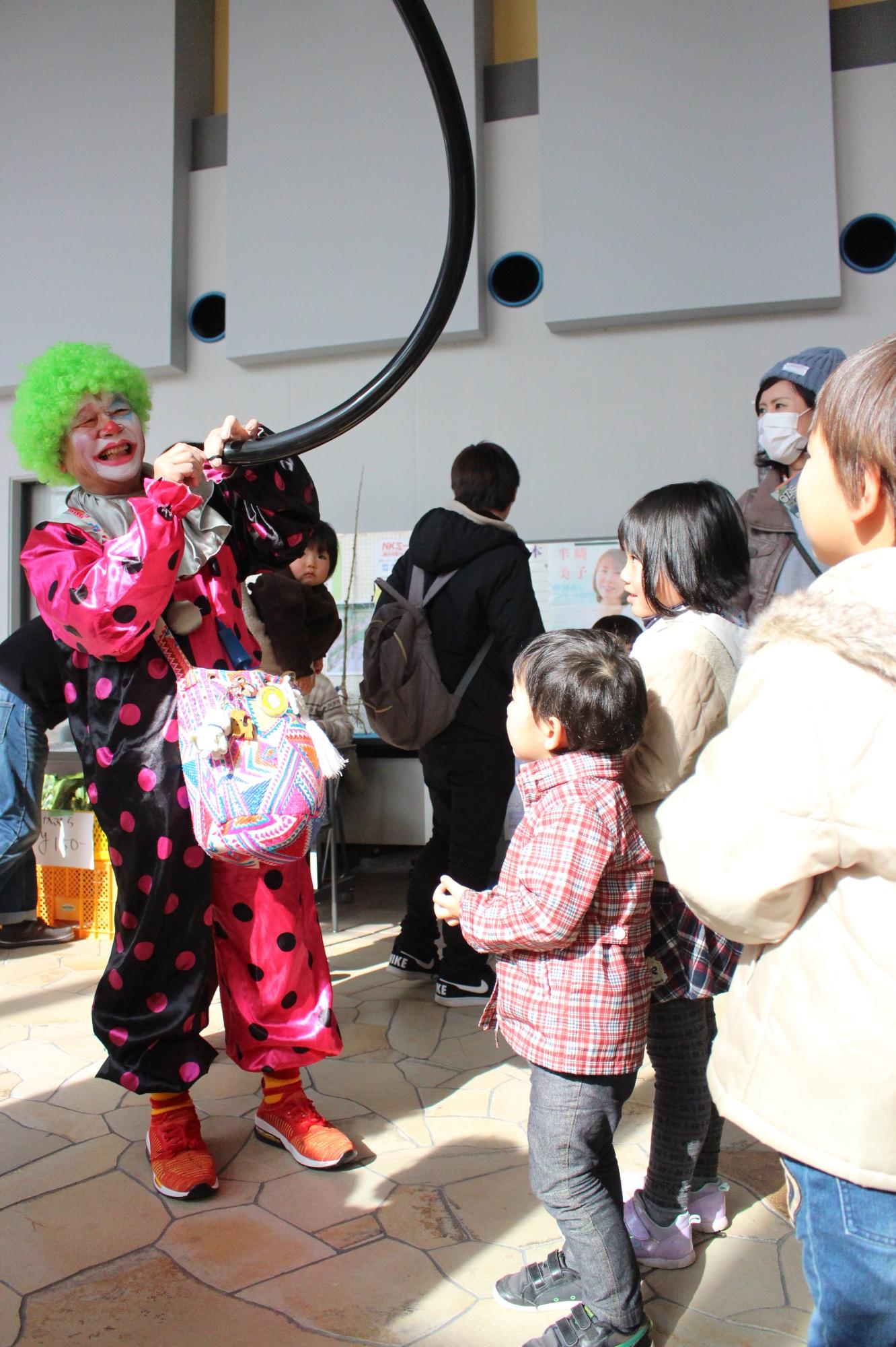 緑のくるくるした頭に黒とピンクの服を着た笑顔のピエロが黒く長い風船を膨らませ、その様子をみる子どもたち