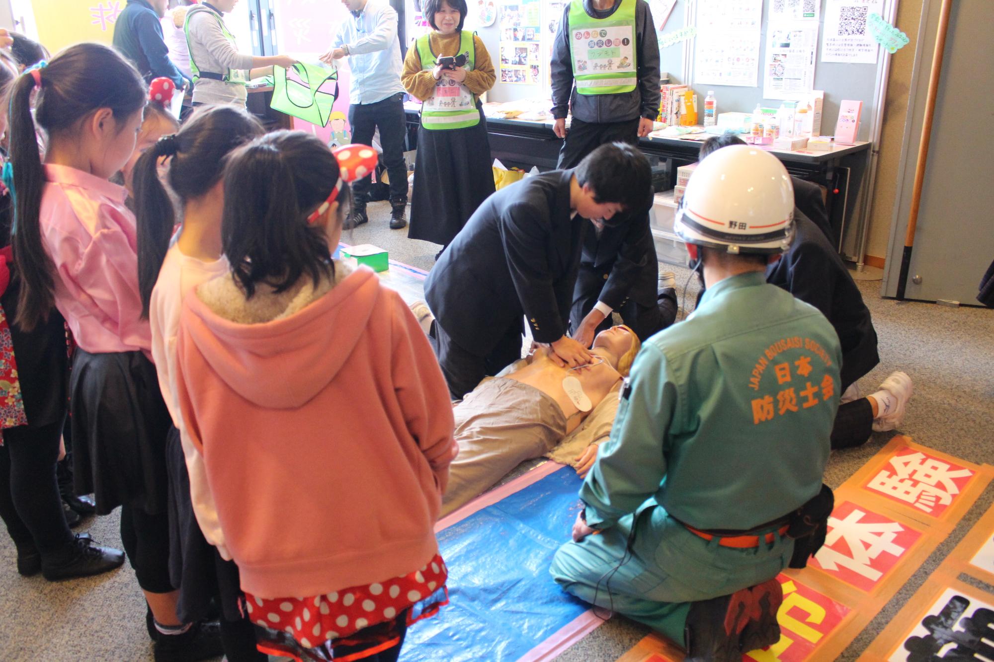 心臓マッサージの体験を行っている男性と、それを見る日本防災士会の方や子どもたち