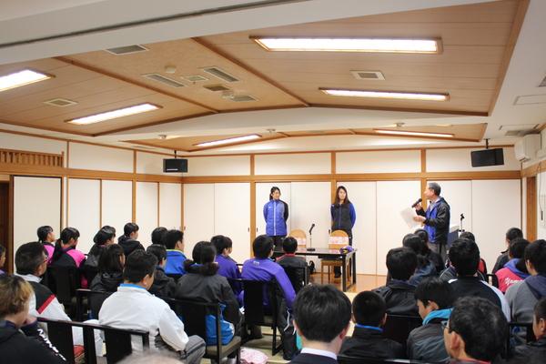 森原さんと深澤さんが代表で前に立ち、司会の男性がお話しして、ジャージを着た人たちが椅子に座り話を聞いている写真