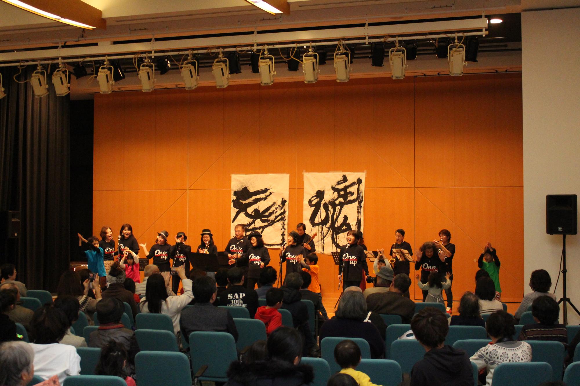 歌を歌ったり、その後ろでギターやトランペットなどを演奏している人や、踊る子どもたちと、それをイスに座って見ている人たちの写真