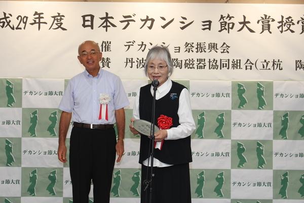 日本デカンショ節大賞授賞した優秀賞 泉 靖子さんがお祝い品を持って挨拶をしている様子を一歩後ろから見ている市長と記念写真